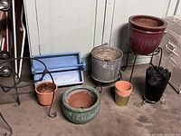 Various plant pots and metal stands arranged on floor, colors include reddish-brown ceramic pot, galvanized metal bucket, green ceramic pot, plastic pots in terracotta and green.