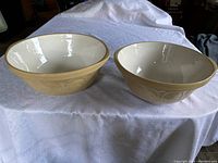 Pair of large beige and white ceramic mixing bowls on white fabric, showing size difference and design pattern on outside rim.