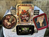 Five vintage metal serving trays displayed on a tiled floor, showing various designs including Olympic Coca-Cola, Samuel Smith's, Pepsi, and vintage artwork of female figures.