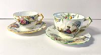 Two vintage Aynsley bone china cups and saucers, showing each set from the side, depicting floral and scenic patterns with gold trim.