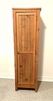 Front view of the wooden storage cupboard showing the entire piece with closed door revealing vertical slat paneling and metal knob.
