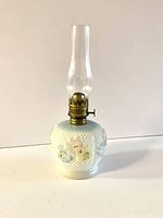 Front view of the Victorian miniature oil lamp showing the white milk glass body with waffle weave texture, blue band at the shoulder, and colorful cosmos flowers. Brass burner and glass chimney in place.