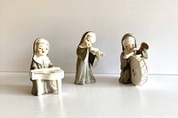 Three porcelain nun figurines arranged in a row, each playing different musical instruments, shown against a neutral background.