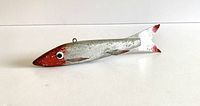 Side view of vintage ice fishing decoy showing silver wooden body, red-colored head and tail tips, metal fins, eye hook on the top.