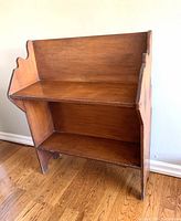 View of the wooden pail bench at an angle showing its front and right side, highlighting the slanted top shelf and lower shelf, and the warm brown wood finish.