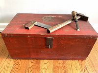 Front view of vintage red painted wooden tool chest with metal hasp, wooden side grips, and old nautical decal on lid. Tools placed on top for scale and context (not included).