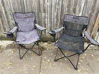 Two black folding camping chairs with mesh seats and cup holders, placed outdoors in front of wooden fence.
