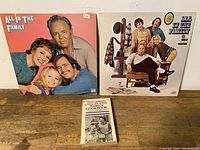Two All In The Family vinyl LP records with cover artwork and the Edith Bunker's cookbook laid out on a wooden surface.