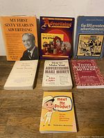 Photo showing all 7 advertising books arranged standing and stacked on a wooden surface, various titles and authors visible.