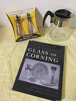 Image of the three items: the packaged Pyrex salt and pepper shakers, the Corning coffee pot carafe with black handle and gold starburst design, and the book titled 'The Complete Cut & Engraved Glass of Corning'.