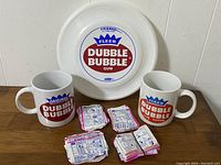 Photo showing two Dubble Bubble mugs, white plastic frisbee with Dubble Bubble Fleer logo, and multiple stacks of folded gum comics on a wooden surface.