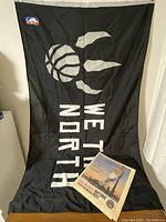 5x3 feet black Raptors flag with claw basketball logo and 'WE THE NORTH' text, NBA patch visible in upper corner, lying flat against floor and wall.