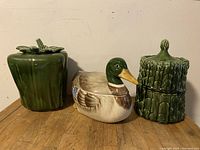 Three vintage ceramic cookie jars on wooden surface: a green bell pepper, a mallard duck, and an asparagus jar.