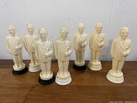 Seven ivory Colonel Harland Sanders figurines in two rows on a wooden surface, all with black bases.