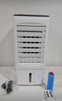 Front and top view of the white portable air cooler with control panel, water level indicator, blue liquid bottle and black attachments shown on a plain background.