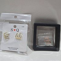 Two pairs of earrings: one pair gold-tone triangular with rhinestones on S Q S925 card in plastic sleeve, one pair rose gold-tone small square earrings in black display box.
