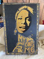 Full view of wood art depicting Nina Simone with gold-painted face and text 'My Baby just Cares For Me' on black background.