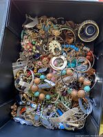 Top-down view of a black box filled with assorted mixed metal and beaded jewellery items including necklaces, bracelets, brooches, and earrings.