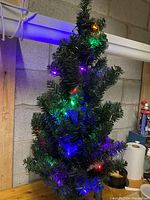 Front and angled view of 3ft artificial Christmas tree with multicolored LED lights, on wooden table against concrete block wall with surrounding storage items.