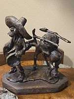 Front and side view of vintage bronze statue depicting a whimsical man with a large hat holding a pan or bowl, standing next to a donkey carrying a load, all mounted on a wooden base.