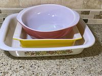 Three vintage baking dishes stacked showing a large rectangular dish with floral pattern, a yellow square dish, and a pink round mixing bowl.