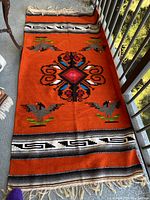 Full view of orange handwoven rug with multicolored geometric and bird motifs and fringed ends, displayed on balcony floor.