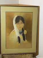 The painting shows a Native girl with closed eyes, traditional attire including feathers and bead necklace, with the artist's signature visible. The piece is framed with a gold frame and pale green mat.