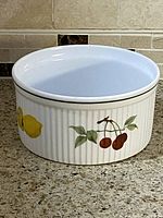 Side angle showing lemon and cherry fruit designs on ribbed white ceramic dish with green rim band.