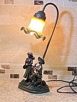Photo of vintage Art Deco table lamp showing the curved black metal arm, glowing yellow ruffled glass shade, and base figurines of boy playing flute and girl holding flower.