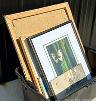Image showing several framed artworks stacked in a black plastic bin, including a visible canvas painting at the back and several glass-framed prints at the front.