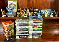 Wide shot showing several Skylanders figures lined up behind stacked Xbox 360 game cases and the Scene It 2 box with mic and cable on table.