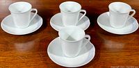 Four white demitasse cups on matching saucers arranged on wooden surface.