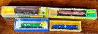 Four N scale model train rail cars in unopened cases arranged on a wooden surface. Visible brands are Bachmann and Minitrix, including Southern Pacific tanker and CP Rail hopper cars.