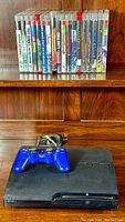 Front overview of PS3 Slim console, blue controller, power cord and row of game cases