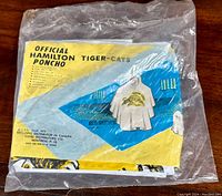 Image showing the vintage Hamilton Tiger-Cats poncho still sealed inside the original clear plastic packaging with product insert visible inside.