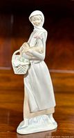 Front view of Lladro porcelain figurine of girl holding cockerel and basket, against wooden background.