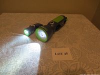Two clip-on flashlights, one smaller and one larger, both black and green with lights on, placed on a lace tablecloth with 'LOT 45' sign