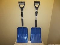 Two blue telescopic emergency snow shovels with black D-shaped handles and padded grips, standing upright against a beige wall.