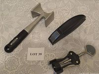 Photo shows three kitchen utensils on white lace fabric: a metal meat tenderizer with black handle, black and metal garlic press, and wing corkscrew with black handles.