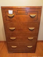 Front view of the wooden dresser showing all five drawers and brass half-moon handles with decorative black inlay on top drawer.