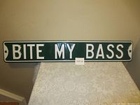 Photo showing a rectangular metal sign with the text "BITE MY BASS" in white embossed letters on a dark green background, placed on a lace cloth against a beige wall.