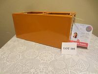Large orange metal storage bin shown from the side on a table with a lace tablecloth. Tagged with Debbie Travis label and labeled LOT 105.