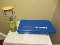 Blue Primus branded portable propane stove in closed position next to the propane lantern on a lace tablecloth.