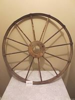Photo of a solid metal wagon wheel with multiple spokes against a beige wall, showing the entire wheel and the table it rests on.