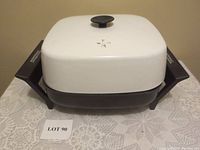 11-inch square electric fry pan with white lid placed on tablecloth. Black side handles visible with 'Toastess' branding.