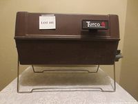 Closed view of brown Turco portable propane BBQ with hinged lid, side handles, and metal stand.