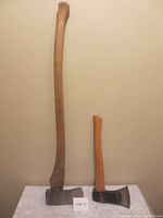 Photo showing two wood chopping axes against a wall, one large (32 inches) and one smaller (15 inches) both with wooden handles and metal heads.