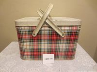 Full view of the exterior of metal picnic basket with red, green, black, and white plaid pattern and two foldable metal handles.