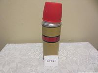 Closed thermos standing upright on a lace tablecloth with a label 'LOT 41' in front, showing the gold finish with red and black stripes and red lid on top.