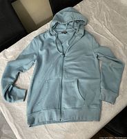 Image showing a laid flat light blue adult zip-up hoodie sweater with a hood, zipper, and front pockets, displayed to show details and condition.